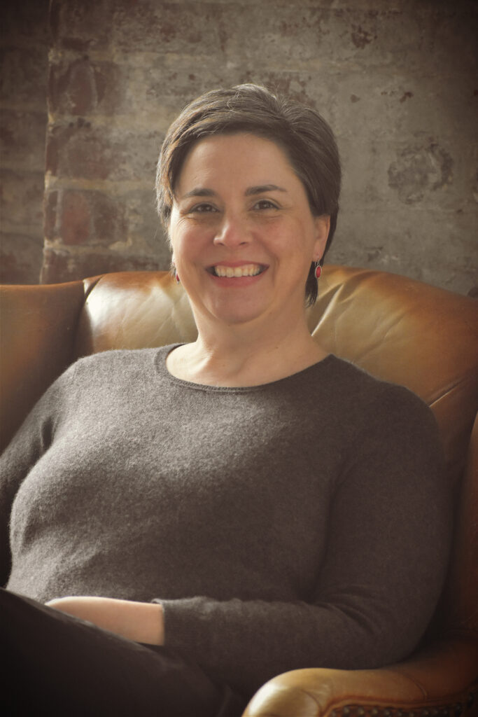 Dr. Jennifer Guiliano seated in a leather chair wearing a grey sweater and black skirt in front of a brick wall.